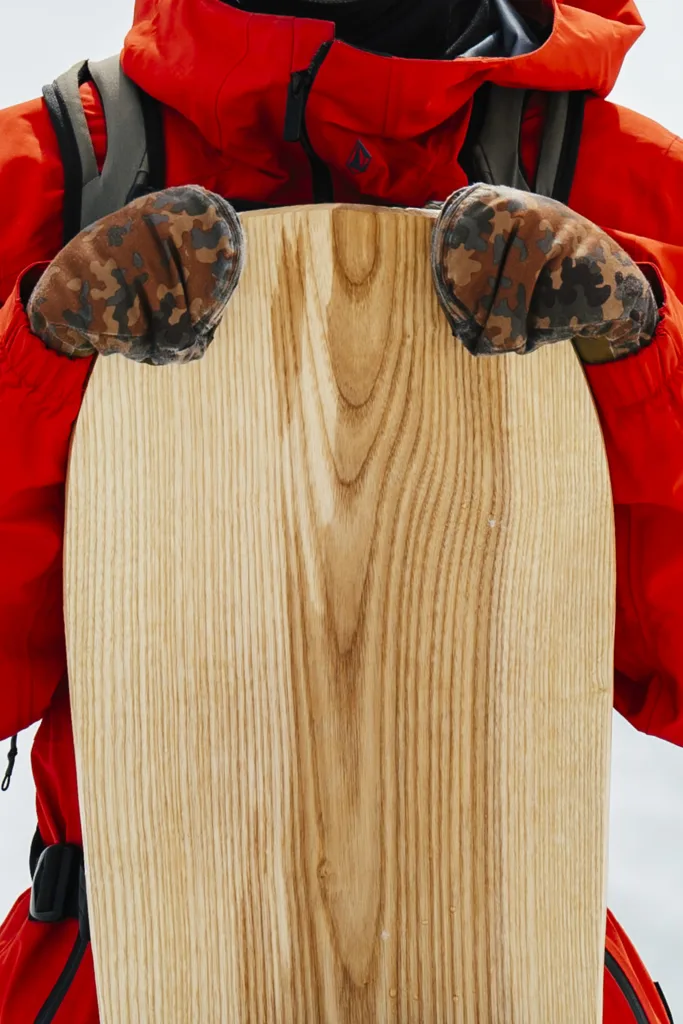 A snowboarder wearing a red jacket is holding a snowboard made completely out of wood.